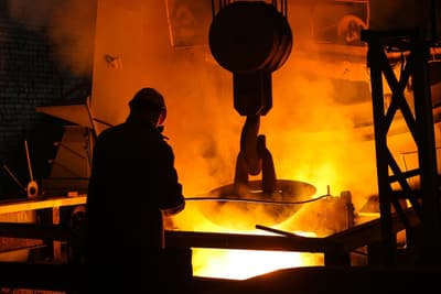 A worker in a helmet at glowing molten steel under a crane hook
